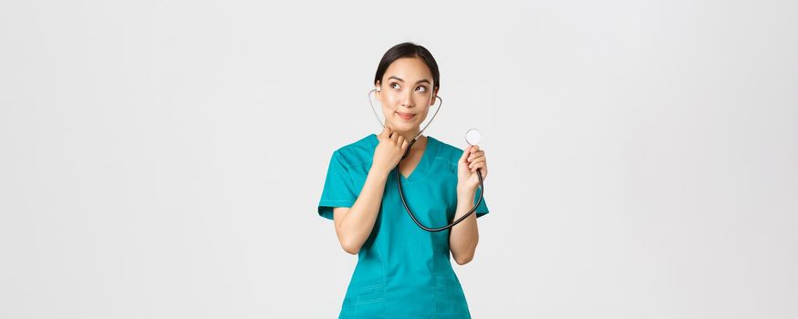 Covid-19, healthcare workers and preventing virus concept. Cute smiling asian female intern learn how listen lungs with stethscope, doctor examine patient, looking thoughtful upper left corner.