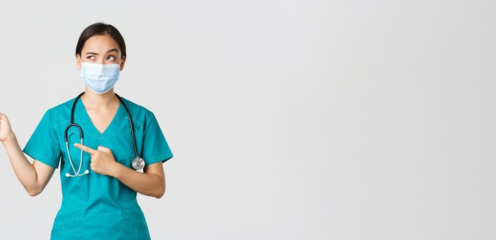 Covid-19, coronavirus disease, healthcare workers concept. Suspicious or doubtful asian female doctor in scrubs and medical mask, looking uncertain and pointing upper left corner, white background.