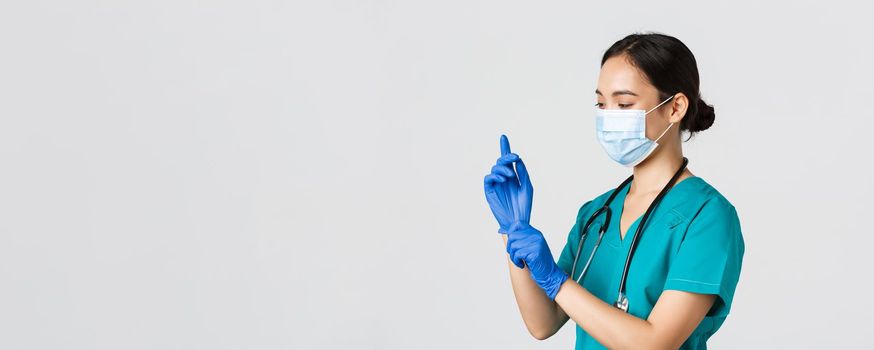 Covid-19, coronavirus disease, healthcare workers concept. Professional smiling asian female nurse, physician in scrubs and medical mask put on rubber gloves for checkup, patient examination.