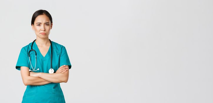 Covid-19, healthcare workers, pandemic concept. Skeptical and reluctant asian female doctor, tired nurse in scrubs cross arms and frowning, smirk displeased, standing white background.
