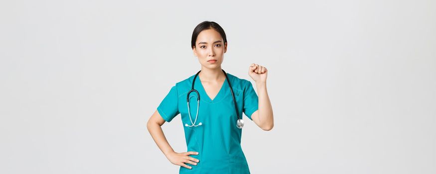 Covid-19, healthcare workers, pandemic concept. Serious-looking confident serious asian female doctor showing support to fellow collegues during coronavirus, raising fist in unity gesture.