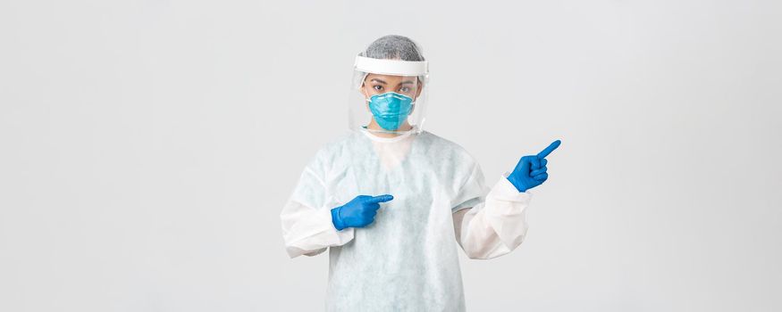 Covid-19, coronavirus disease, healthcare workers concept. Professional female doctor, tech lab employee in personal protective equipment pointing fingers right, showing way, white background.