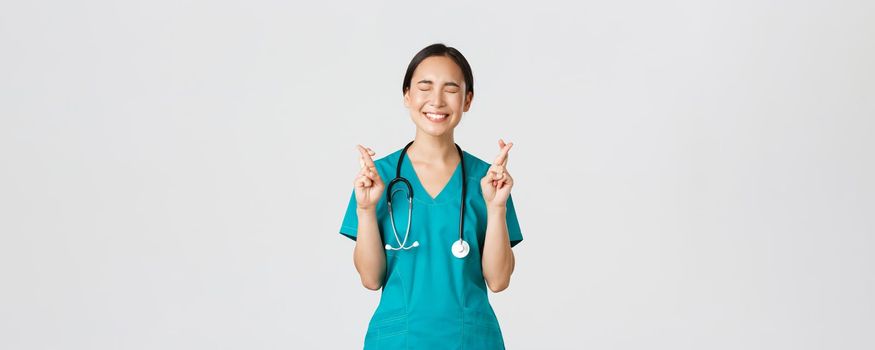 Covid-19, healthcare workers, pandemic concept. Optimistic happy and hopeful asian female nurse, intern waiting for promotion, cross fingers good luck and close eyes, smiling excited.