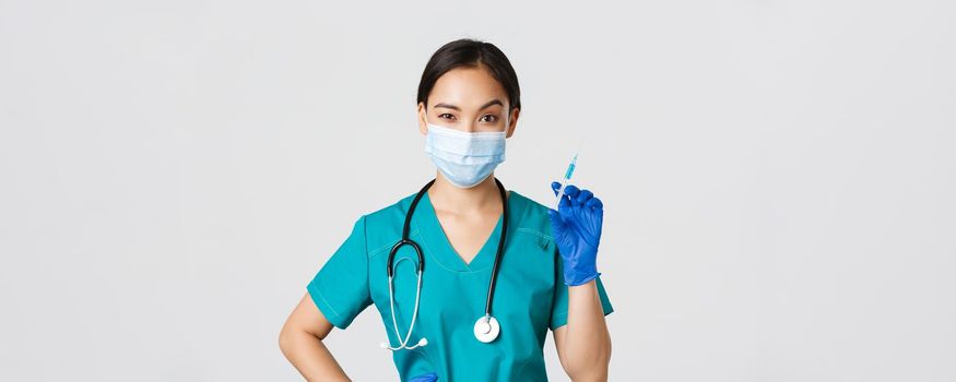 Covid-19, coronavirus disease, healthcare workers concept. Sassy cute asian female nurse, intern in medical mask and gloves holding syringe with vaccine, ready to make shot, white background.