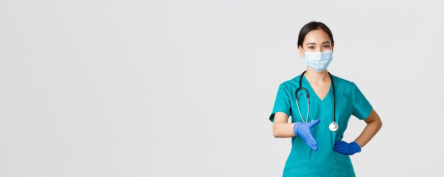 Covid-19, coronavirus disease, healthcare workers concept. Confident young asian female doctor, physician in scrubs and medical mask, gloves, extend hand for handshake, greeting patient.