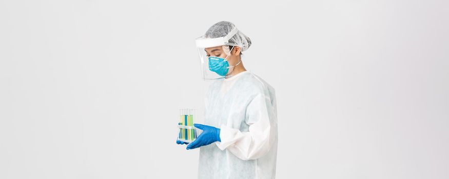 Covid-19, coronavirus disease, healthcare workers concept. Side view of careful serious asian female doctor, tech lab employee in personal protective equipment holding test-tube probes of vaccines.