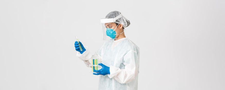 Covid-19, coronavirus disease, healthcare workers concept. Side view of careful serious asian female doctor, tech lab employee in personal protective equipment holding test-tube probes of vaccines.