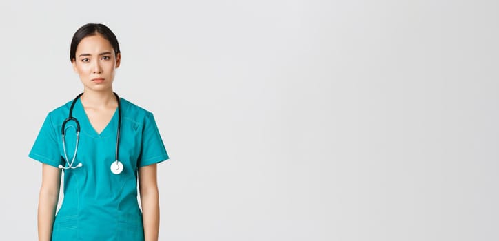 Covid-19, healthcare workers, pandemic concept. Exhausted young asian female nurse, doctor looking tired after shift in hospital, looking sad with fatigue, standing white background in scrubs.
