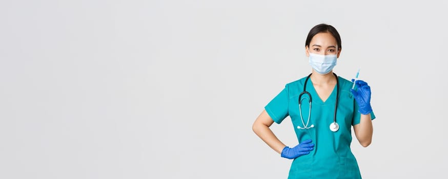 Covid-19, coronavirus disease, healthcare workers concept. Smiling confident asian female nurse in scrubs and medical mask, holding syringe with vaccine, doing vaccination shot, white background.