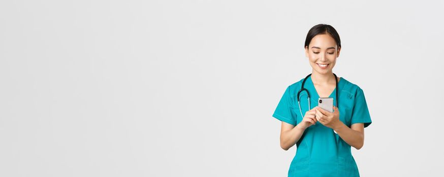 Covid-19, healthcare workers and preventing virus concept. Smiling happy beautiful asian female intern, doctor making phone call, looking at mobile phone screen pleased, messaging or using app.