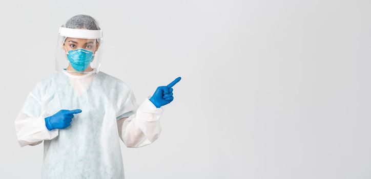 Covid-19, coronavirus disease, healthcare workers concept. Professional female doctor, tech lab employee in personal protective equipment pointing fingers right, showing way, white background.