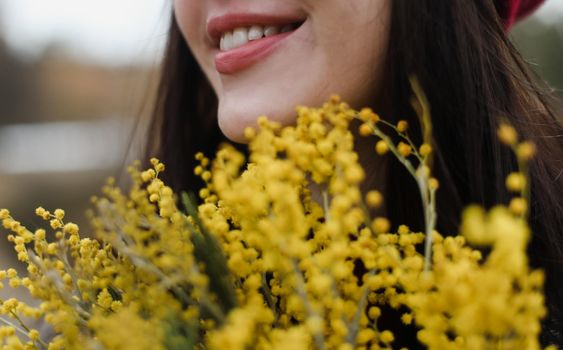 close-up portrait of a smiling brunette girl with red lips enjoys a large bouquet of fresh beautiful yellow mimosa bouquet outdoors