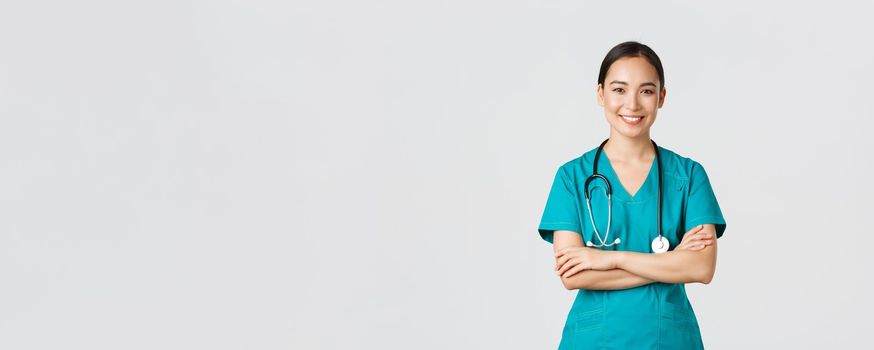 Covid-19, healthcare workers, pandemic concept. Portrait of confident smiling, attractive asian female nurse in scrubs, with stethoscope, cross arms chest and looking at camera, white background.