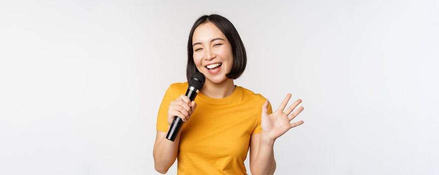 Happy asian girl dancing and singing karaoke, holding microphone in hand, having fun, standing over white background.