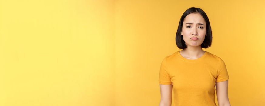Disappointed asian girl sulking, looking upset, feel unair, standing in yellow t-shirt over white background. Copy space