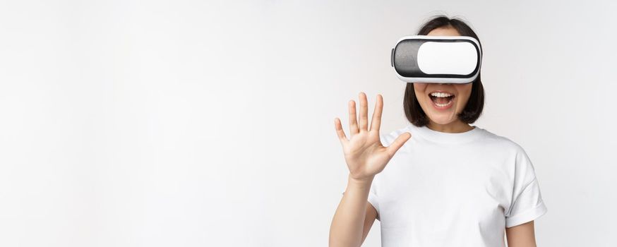 Happy asian woman using VR headset, waving raised hands and laughing, using virtual reality glasses, standing over white background.