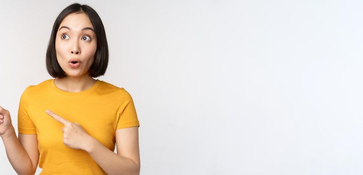 Surprised asian woman pointing and looking left at copy space, promo sale, showing advertisement with impressed face expression, standing over white backround.