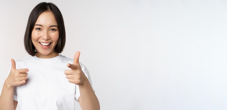 Image of smiling asian girl pointing fingers at camera, choosing, inviting you, congratulating, standing in tshirt over white background.