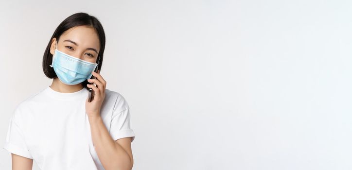 Health and covid-19 concept. Smiling asian girl in medical face mask talking on phone, answer mobile call, standing over white background.