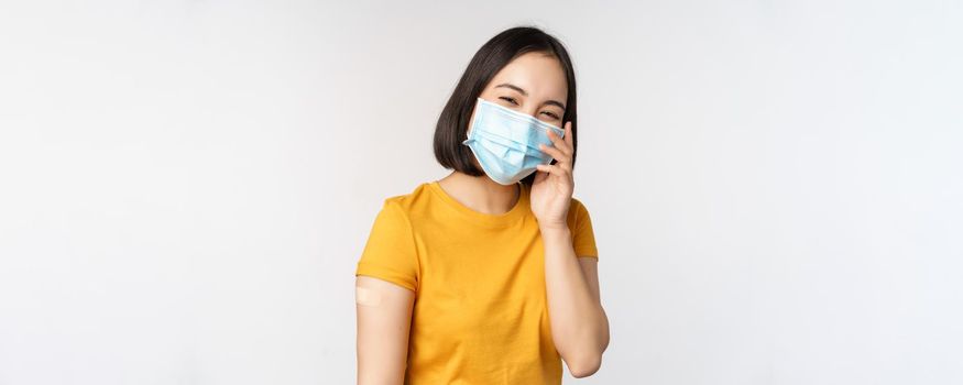 Covid-19, vaccination and healthcare concept. Cute asian girl in medical face mask, showing band aid after coronavirus vaccination, standing over white background.