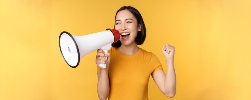 Announcement. Happy asian woman shouting loud at megaphone, recruiting, protesting with speaker in hands, standing over yellow background.