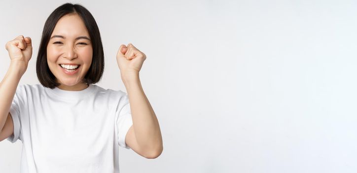 Portrait of enthusiastic asian woman winning, celebrating and triumphing, raising hands up, achieve goal or success, standing over white background.