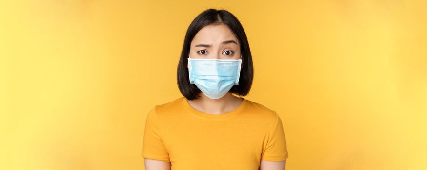 Portrait of skeptical and confused asian woman in medical face mask, raising eyebrow doubtful, standing over yellow background.