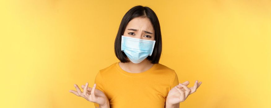 Close up portrait of confused asian woman in medical face mask, shrugging shoulders and looking puzzled, standing against yellow background.