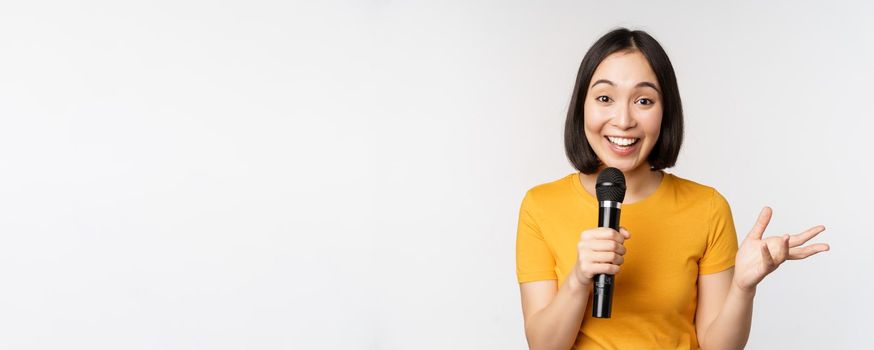 Image of young asian woman talking in microphone, perfom with mic, giving speech, standing in yellow tshirt against white background.