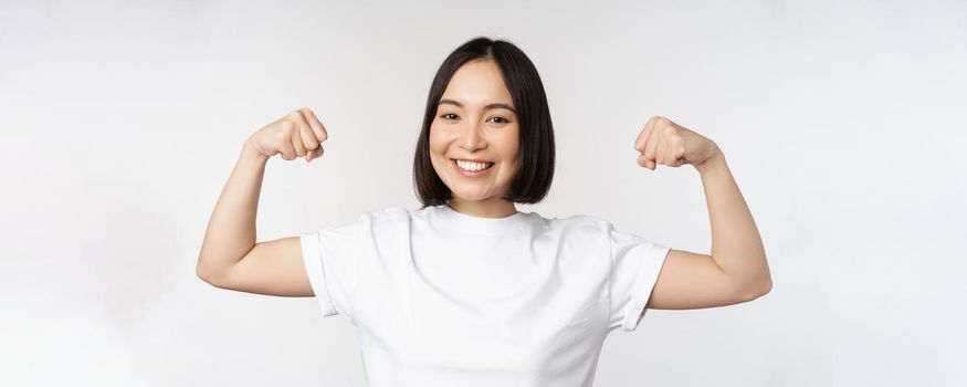 Smiling asian woman showing flexing biceps, muscles strong arms gesture, standing in white tshirt over white background.