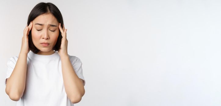 Portrait of asian girl feeling headache, migraine or being ill, standing in white t-shirt over white background. Copy space