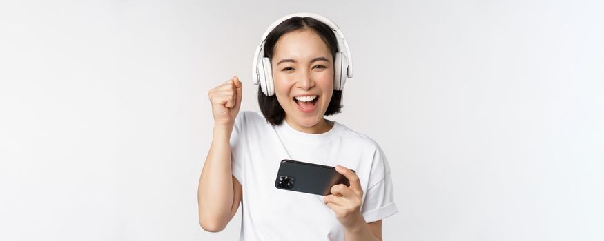 Happy asian girl gamer, playing on mobile phone, watching on smartphone, wearing headphones, standing over white background.