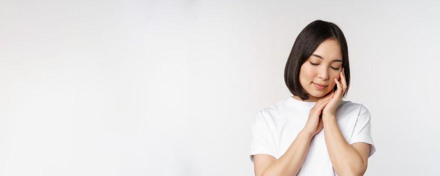 Portrait of tender and feminine young asian woman, looking down, gently touching face, concept of skin care and cosmetology, white background.
