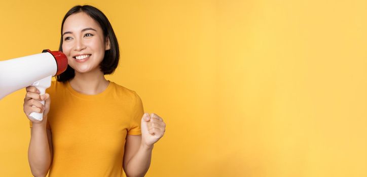 Smiling asian woman standing with megaphone, announcing smth, advertising product, standing over yellow background.