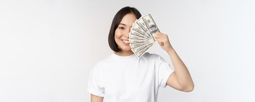 Portrait of smiling asian woman holding dollars money, concept of microcredit, finance and cash, standing over white background.