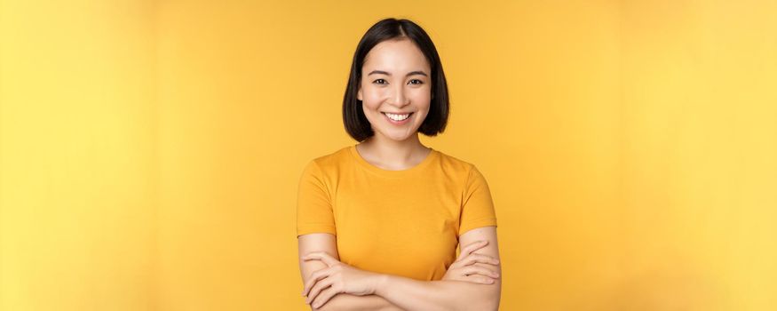 Confident asian girl cross arms on chest, smiling and looking assertive, standing over yellow background.