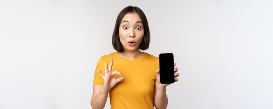 Excited asian girl showing mobile phone screen, okay sign, recommending smartphone app, standing in yellow tshirt over white background.