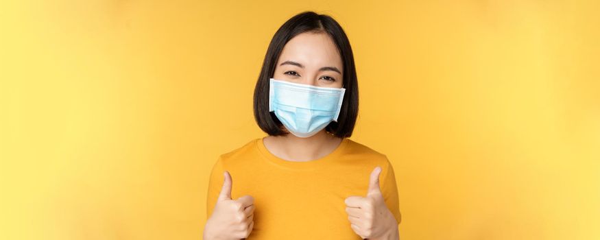 Cheerful korean woman in medical face mask, support people during pandemic, wear personal protective equipment from covid-19, showing thumbs up in approval, yellow background.