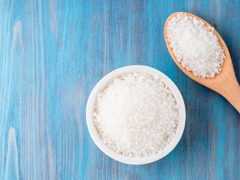 White bowl with large sea salt and wooden spoon on blue bright wooden table. Top view.