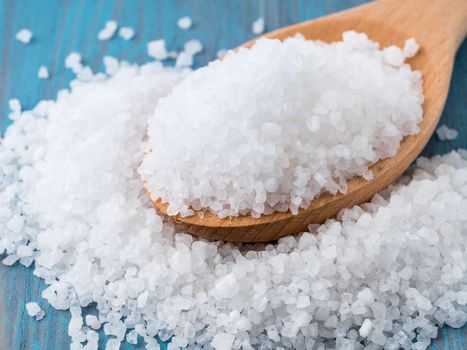 large wooden spoon with big sea salt for the bright blue wooden background, on the table scattered salt crystals, side view