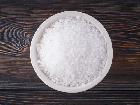 White bowl with large sea salt on brown bright dark wooden table. Top view.