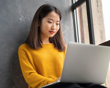 Asian woman working on laptop at home or in cafe on window. Young female in bright yellow jumper is sitting and typing on computer. Business oriental lady in windowsill