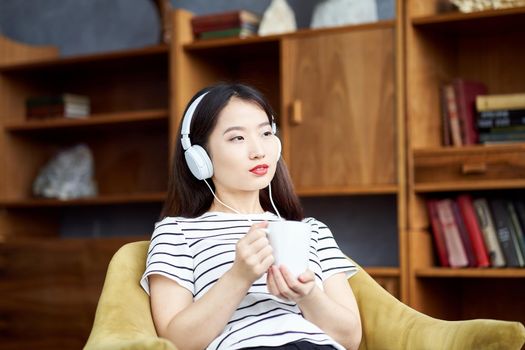 Young beautiful Asian girl listening to music with headphones sitting in chair and looking out window. Woman drinking coffee from mug. Morning routine and contemplation