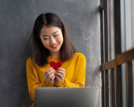 Online Dating on Valentines day at distance during quarantine and self-isolation. Woman showing red heart symbol of love in front of laptop and talking on video call with man