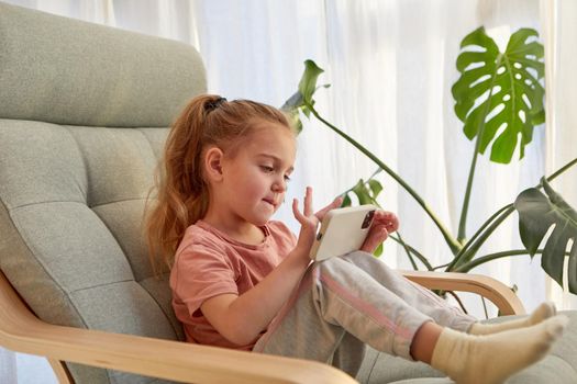 Side view of cute little girl with long blond hair in casual clothes playing video game on mobile phone while sitting on comfortable armchair in daylight at home