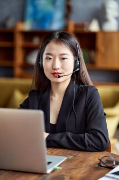 Happy asian businesswoman in headset speaking by conference call and video chat on laptop in office, smiling chinese support service agent consulting customer. Female portrait looking at camera