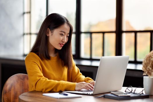 Asian woman working on laptop at home or in cafe. Young lady in bright yellow jumper is sitting at desk typing on computer. Business oriental female in front of window