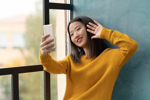 Young Asian woman takeing selfie and waving hand. Beautiful teenage girl with bright red lips siting on window and communicating via video call with boyfriend