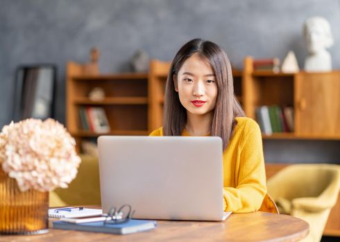 Asian woman working on laptop at home or in cafe. Young lady in bright yellow jumper is sitting at desk typing on computer. Business oriental female in front of window