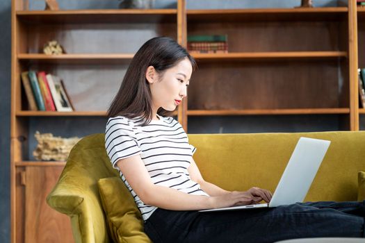 Asian woman looking at laptop, working at home, liying on sofa. Beautiful Japanese female freelancer smiling and typing at computer in hotel on couch